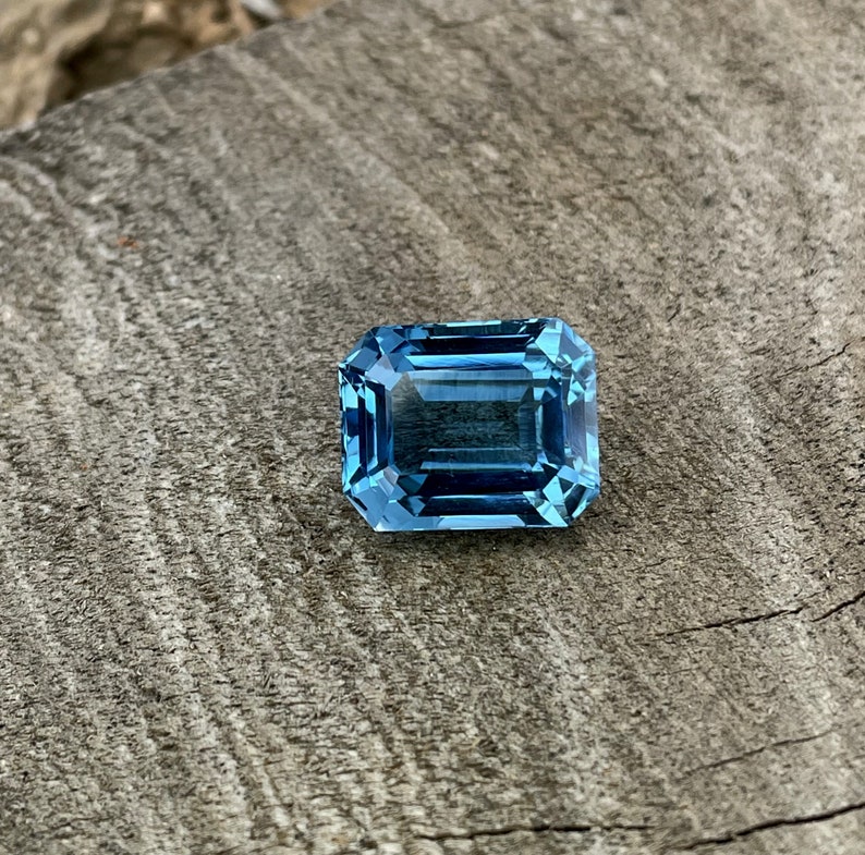 May include: A rectangular, light blue gemstone with an emerald cut, set against a weathered, grey wooden surface. The facets of the gem reflect light, creating a sparkling effect. The stone's colour is a vivid, clear blue, reminiscent of the sea.