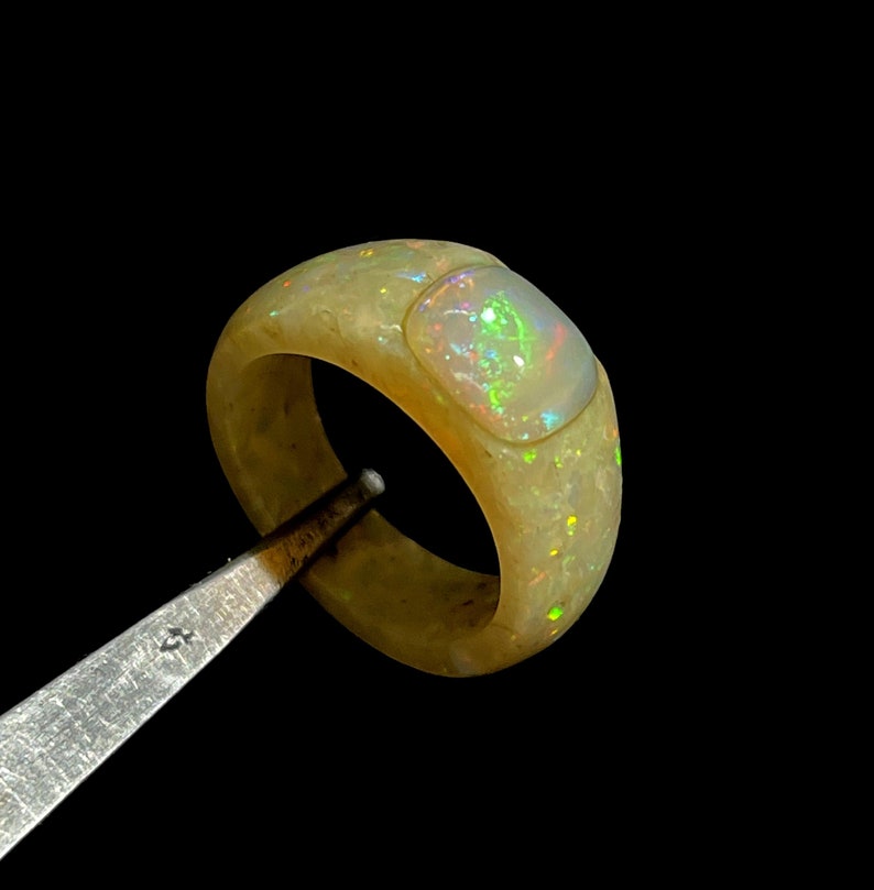 May include: A wide, light-colored opal ring with a rectangular opal inlay. The ring has a smooth, polished surface and displays flashes of green, blue, and yellow. The ring is held by a metal tool against a black background.