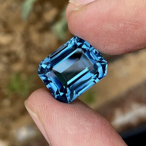 May include: A rectangular, faceted gemstone in a deep blue shade. The gem is held between two fingers, highlighting its clarity and cut. The facets reflect light, creating a brilliant display. The gem's colour varies from light to dark blue.