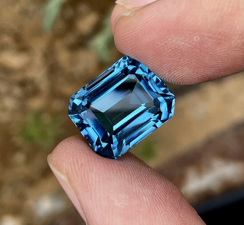 May include: A rectangular, faceted gemstone in a deep blue shade. The gem is held between two fingers, highlighting its clarity and cut. The facets reflect light, creating a brilliant display. The gem's colour varies from light to dark blue.