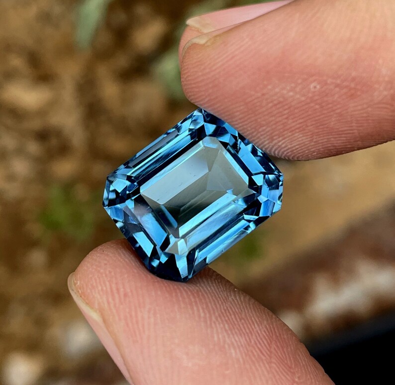 May include: A rectangular-cut gemstone in a deep blue shade. The gem is held between two fingers, highlighting its clarity and sparkle. The cut of the stone creates multiple facets, reflecting light. A beautiful piece of jewellery, perfect for a ring or pendant.
