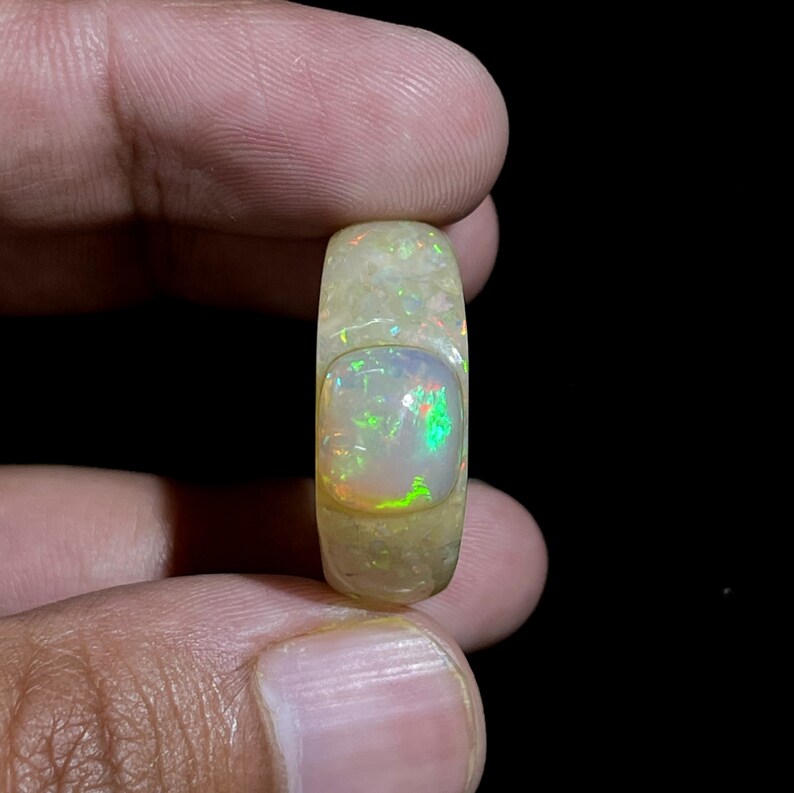 May include: A polished opal gemstone ring with a rectangular center stone. The opal displays a play of color with flashes of green, red, and yellow. The ring has a light yellow hue and is held by a person's fingers against a black background.