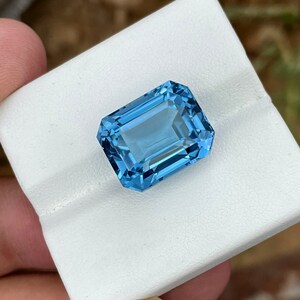 May include: A rectangular, faceted, light blue gemstone. The gem is held on a white surface. The gem has a bright, clear appearance, with multiple facets reflecting light. The colour is a vibrant, sky blue.