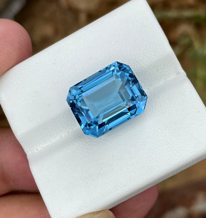 May include: A rectangular, faceted, light blue gemstone. The gem is held on a white surface. The gem has a bright, clear appearance, with multiple facets reflecting light. The colour is a vibrant, sky blue.