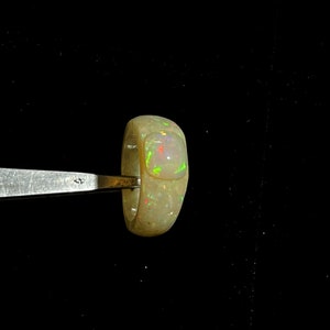 May include: A light-colored opal ring with a rectangular, raised stone. The opal displays flashes of red, green, and yellow. The ring is held by a metal tool against a black background. The ring's band is thick and rounded.