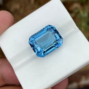 May include: A rectangular, faceted gemstone in a vibrant, light blue colour. The gem is set against a white background, highlighting its clarity and brilliance. The cut of the stone allows light to reflect, creating a sparkling effect. This gemstone is likely for jewellery.