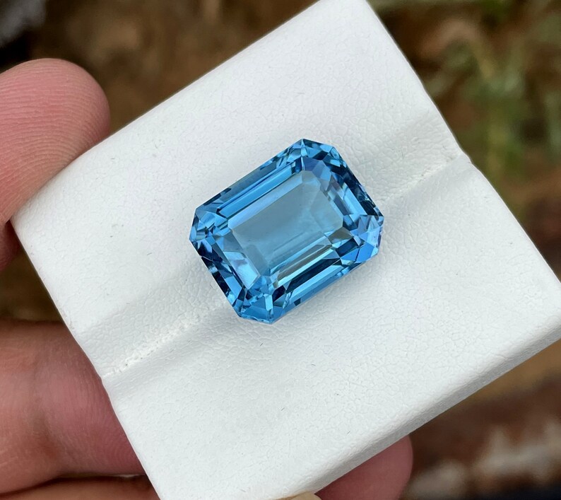 May include: A rectangular, faceted gemstone in a vibrant, light blue colour. The gem is set against a white background, highlighting its clarity and brilliance. The cut of the stone allows light to reflect, creating a sparkling effect. This gemstone is likely for jewellery.