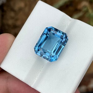 May include: A rectangular, light blue gemstone with a step cut. The gem is held on a white surface, showcasing its clarity and facets. The stone's colour is a vibrant, clear blue, reflecting light.