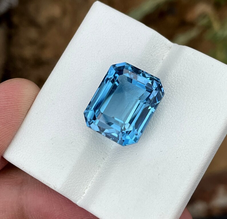 May include: A rectangular, light blue gemstone with a step cut. The gem is held on a white surface, showcasing its clarity and facets. The stone's colour is a vibrant, clear blue, reflecting light.