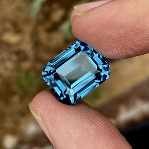 May include: A rectangular-cut gemstone in a striking, transparent blue. The gem is held between two fingers, highlighting its detailed facets and reflective qualities. The facets of the gem create a sparkling effect as they catch the light.