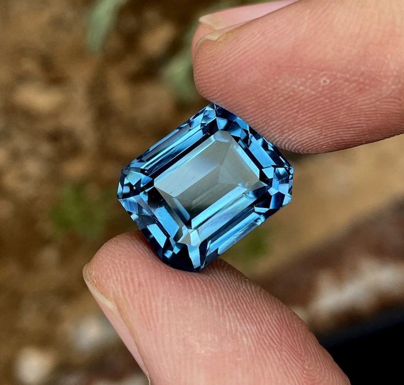 May include: A rectangular-cut gemstone in a striking, transparent blue. The gem is held between two fingers, highlighting its detailed facets and reflective qualities. The facets of the gem create a sparkling effect as they catch the light.