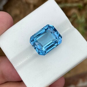 May include: A rectangular, light blue gemstone with multiple facets. The gem is displayed on a white surface, highlighting its clarity and sparkle. The facets catch the light, creating a brilliant effect. The overall appearance suggests a high-quality precious stone.