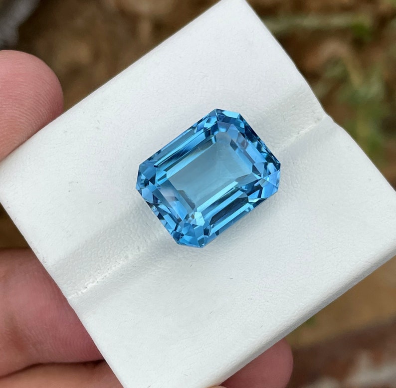 May include: A rectangular, light blue gemstone with multiple facets. The gem is displayed on a white surface, highlighting its clarity and sparkle. The facets catch the light, creating a brilliant effect. The overall appearance suggests a high-quality precious stone.