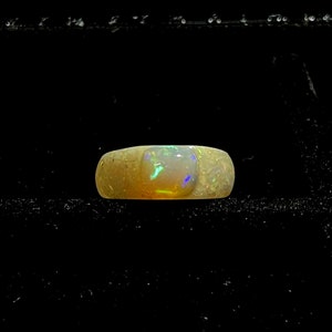 May include: A wide, textured ring with a large, rectangular opal stone. The opal displays a vibrant play of color, including green, blue, and yellow hues. The ring's band has a light, speckled appearance, and the background is black.
