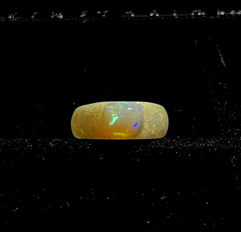May include: A wide, textured ring with a large, rectangular opal stone. The opal displays a vibrant play of color, including green, blue, and yellow hues. The ring's band has a light, speckled appearance, and the background is black.