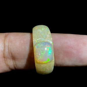 May include: A light-colored opal ring with a rectangular opal stone. The ring has a textured surface and a bright, iridescent display of colors within the opal, including green, orange, and blue hues. The ring is held by a finger against a black background.