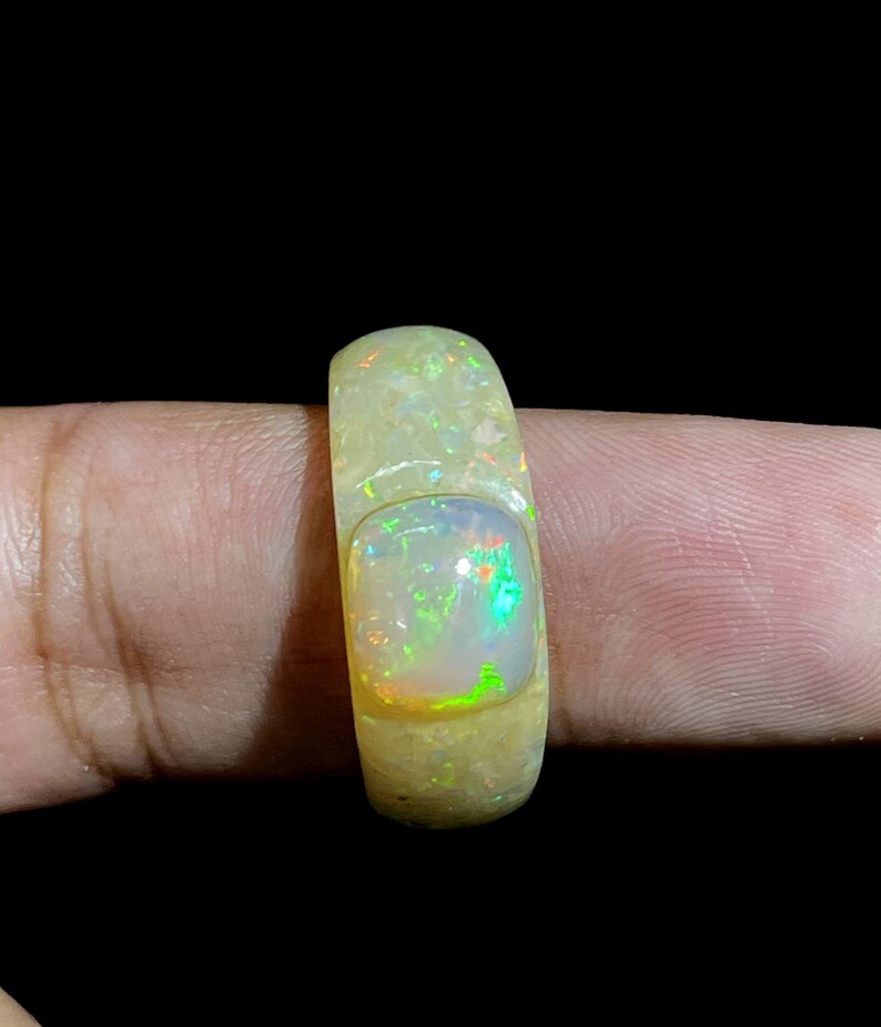 May include: A light-colored opal ring with a rectangular opal stone. The ring has a textured surface and a bright, iridescent display of colors within the opal, including green, orange, and blue hues. The ring is held by a finger against a black background.