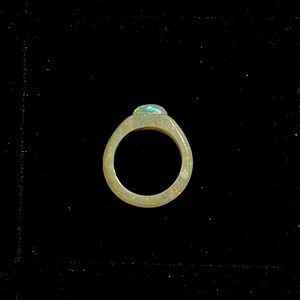 May include: A ring with a light-colored band and a large, oval-shaped opal gemstone. The opal displays a play of color with flashes of green, blue, and red. The ring is set against a black background.