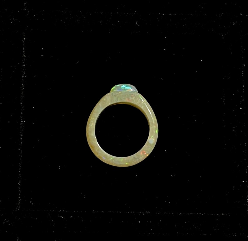 May include: A ring with a light-colored band and a large, oval-shaped opal gemstone. The opal displays a play of color with flashes of green, blue, and red. The ring is set against a black background.