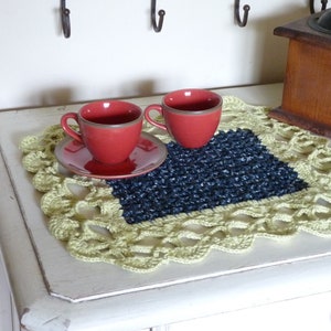May include: Two red ceramic coffee cups with saucers on a crocheted placemat with a blue center. The placemat is on a white table.