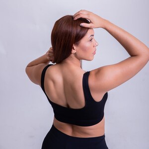 May include: A woman wearing a black sports bra and black leggings, with her hair pulled back, is standing with her back to the camera. She is looking over her shoulder to the right.