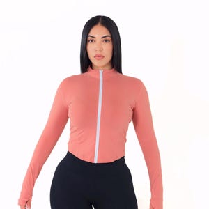 May include: A woman wearing a long-sleeved, salmon-colored zip-up top with a white zipper and black leggings.