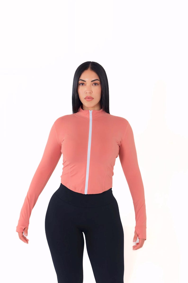 May include: A woman wearing a long-sleeved, salmon-colored zip-up top with a white zipper and black leggings.