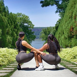 May include: Two people wearing black athletic wear are doing squats facing away from the camera. They are on a stone path with green grass and trees on either side. The path leads to a view of a river and a hillside in the distance.