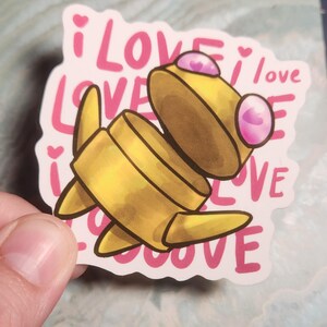 May include: A yellow cartoon character sticker with pink accents and the words "i love" repeated in a pink font. The sticker is held by a hand.