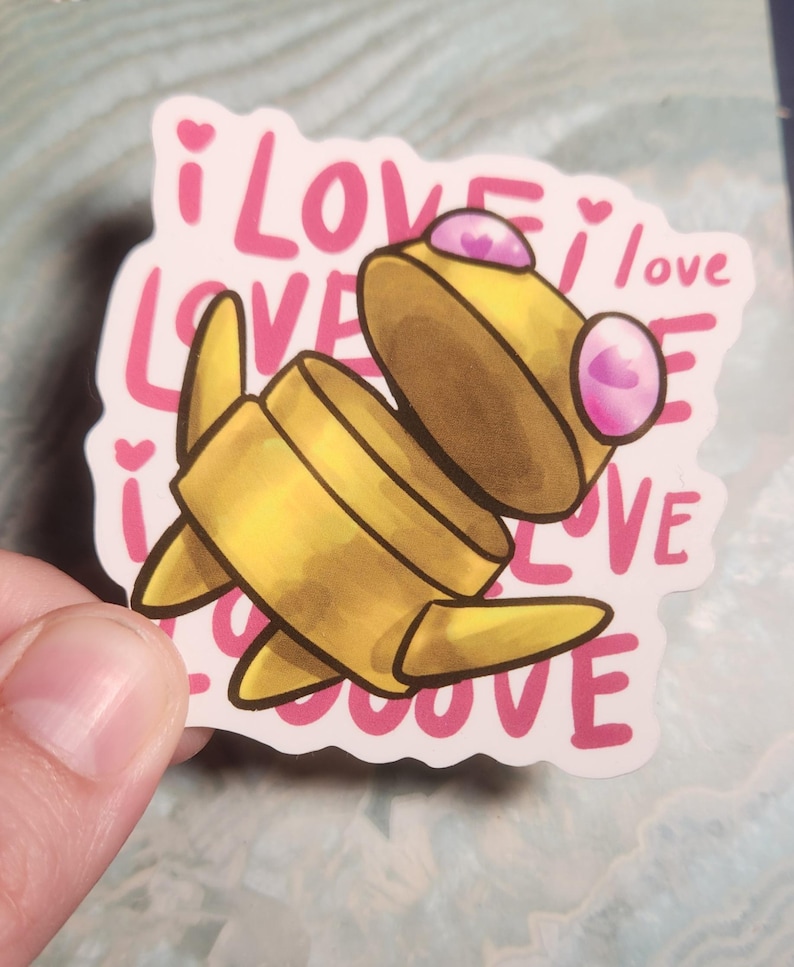 May include: A yellow cartoon character sticker with pink accents and the words "i love" repeated in a pink font. The sticker is held by a hand.