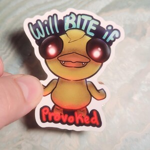 May include: A sticker featuring a cartoon duck with glowing red eyes and a menacing expression. The text "Will BITE if Provoked" is written above and below the duck in a playful font. The duck is yellow with orange accents.