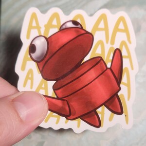 May include: A red cartoon sticker of a creature with large white eyes and the word "AAAA" repeated in a yellow font. The sticker is held by a hand.