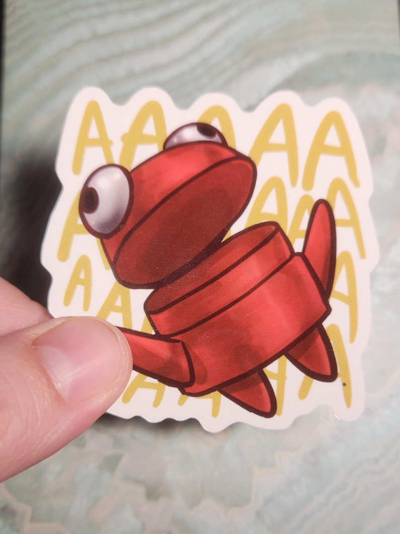 May include: A red cartoon sticker of a creature with large white eyes and the word "AAAA" repeated in a yellow font. The sticker is held by a hand.