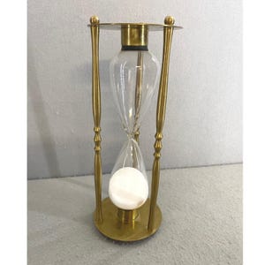 May include: An hourglass featuring a polished brass frame with a circular base and top. The clear glass bulb contains white sand, which is currently gathered in the bottom section. The structure is supported by three slender, vertical brass pillars.