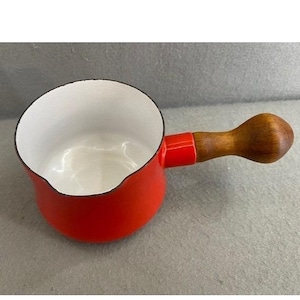 May include: A small, red enamel saucepan with a white interior and a wooden handle. The handle is attached to the pot with a red connector. The saucepan is designed for cooking and has a classic, vintage aesthetic.