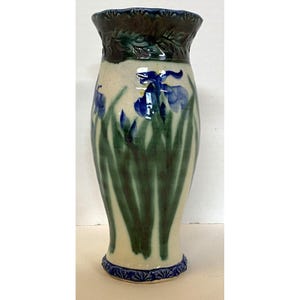May include: A ceramic vase with a cream-colored body, featuring a hand-painted design of blue irises and green leaves. The rim and base have decorative blue and brown accents. The vase is approximately 10 inches tall, with a flared top and a slightly tapered body.