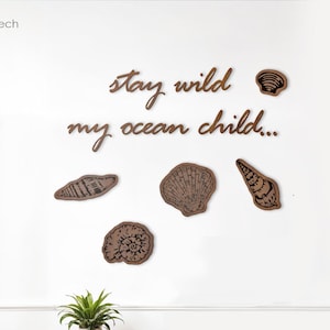 以下が含まれることがあります： 「stay wild my ocean child...」というテキストと、装飾的な貝殻5つが付いた木製の壁飾り。装飾はリビングルームの白い壁に取り付けられています。
