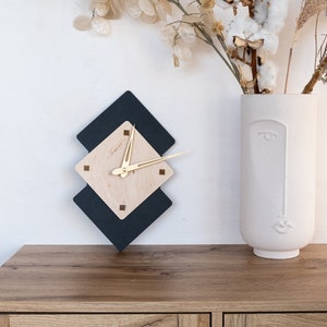 May include: A modern wall clock with a geometric design. The clock features a light wood center with gold hands, set against a black diamond shape. The clock is mounted on a white wall above a wooden cabinet. A white vase with a face design holds dried flowers.