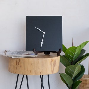 Puede incluir: Un reloj cuadrado negro con manecillas blancas sobre una mesa de madera con una planta verde en el fondo.