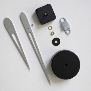 May include: Clock-making components arranged on a white surface. The parts include two silver clock hands, a black clock motor, a black circular base, and various small metal pieces. The hands are approximately 10 inches long.