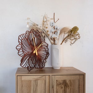 May include: A wooden wall clock with a brown floral design. The clock has gold hands and is set against a white wall with a vase of dried flowers.