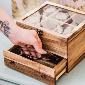 Pu&ograve; includere: Una scatola di legno con un coperchio di vetro e un cassetto. La scatola ha pi&ugrave; scomparti per riporre piccoli oggetti, come gioielli o orologi. Il cassetto &egrave; aperto e contiene un paio di occhiali.