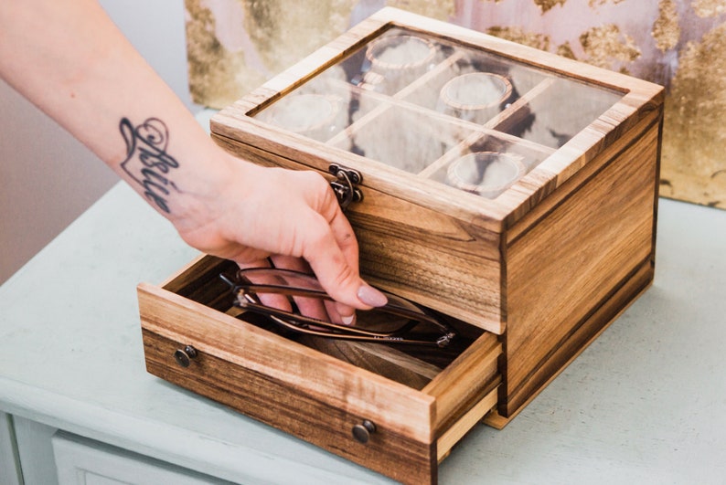 Pu&ograve; includere: Una scatola di legno con un coperchio di vetro e un cassetto. La scatola ha pi&ugrave; scomparti per riporre piccoli oggetti, come gioielli o orologi. Il cassetto &egrave; aperto e contiene un paio di occhiali.