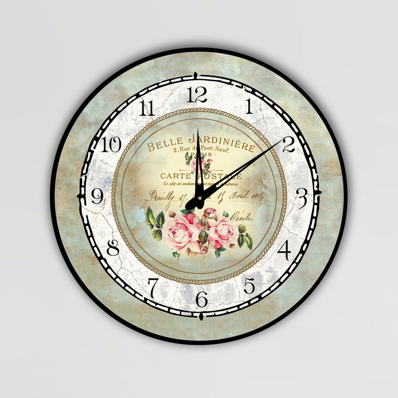 Pu&ograve; includere: Orologio da parete rotondo con un design vintage invecchiato. Il quadrante presenta una composizione floreale di rose rosa, il testo "BELLE JARDINIERE" e lancette nere. L'orologio ha una cornice nera e uno sfondo azzurro e crema.