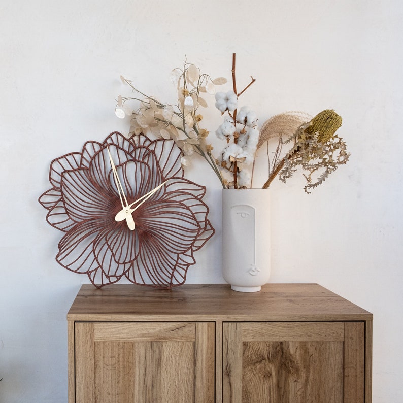 May include: A brown, decorative wall clock shaped like a flower with gold hands. The clock is hanging on a white wall next to a vase with dried flowers.