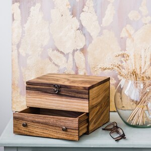 Pu&ograve; includere: Una scatola di legno con un cassetto e un coperchio incernierato. La scatola &egrave; in legno scuro con una venatura naturale. Il cassetto &egrave; aperto e il coperchio &egrave; chiuso. La scatola &egrave; appoggiata su un tavolo blu chiaro.