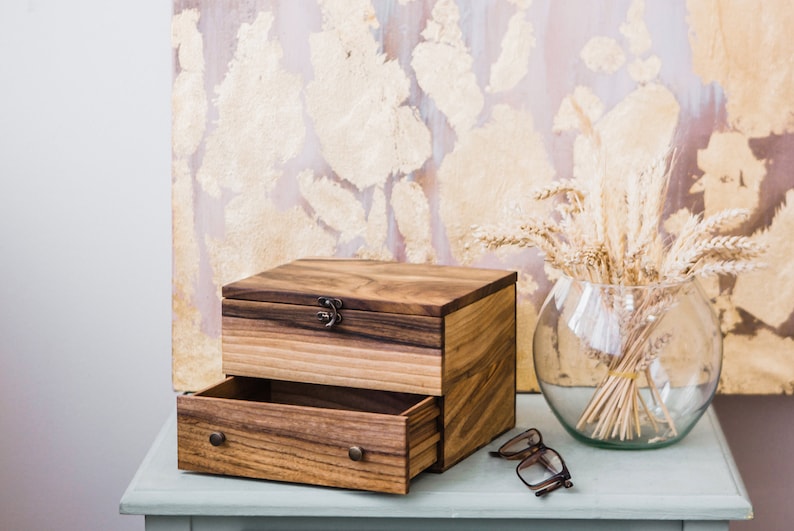 Pu&ograve; includere: Una scatola di legno con un cassetto e un coperchio incernierato. La scatola &egrave; in legno scuro con una venatura naturale. Il cassetto &egrave; aperto e il coperchio &egrave; chiuso. La scatola &egrave; appoggiata su un tavolo blu chiaro.