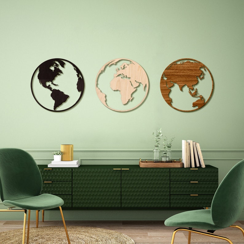 May include: Three wooden wall decor pieces shaped like the Earth, each with a different continent highlighted. The continents are painted in black, brown, and natural wood tones. The decor pieces are hanging on a light green wall above a dark green dresser with gold handles.