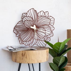 May include: A brown floral-shaped wall clock with white hands. The clock is sitting on a wooden table with a green plant in the background.