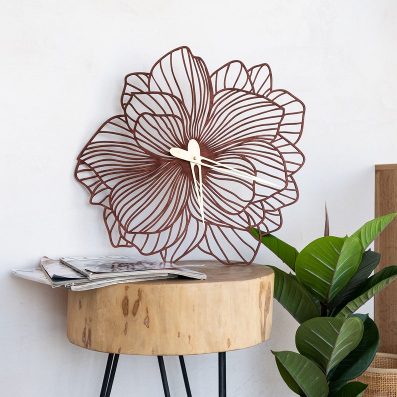 May include: A brown floral-shaped wall clock with white hands. The clock is sitting on a wooden table with a green plant in the background.